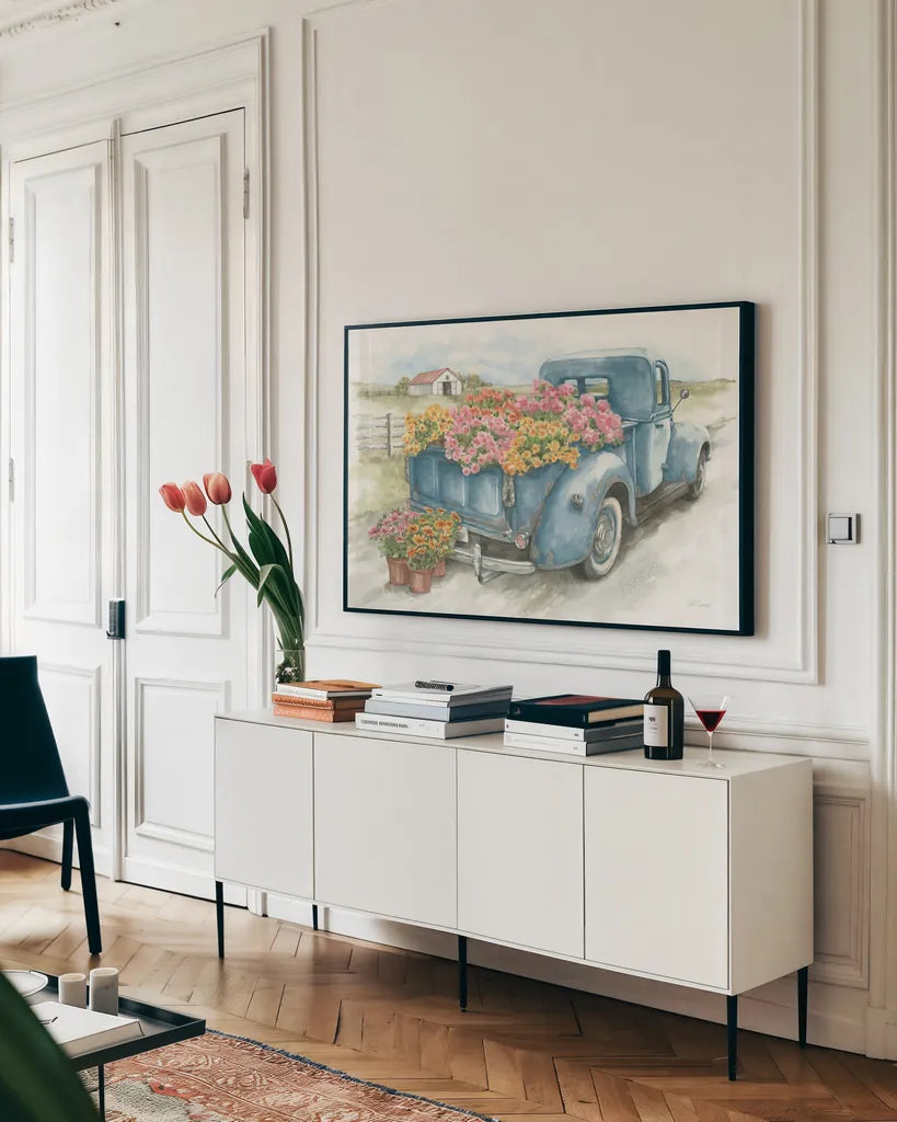 Truck with Flowers on Blue by Leslie Trimbach. shown as a framed stretched canvas in a living room above a console. Available as a archival giclee rolled canvas, fine art paper, or poster print. SKU 86547i.